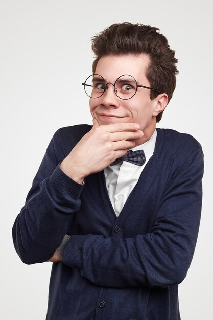 Funny pondering young man in elegant clothes and eyeglasses rubbing chin and looking at camera with smile against white background