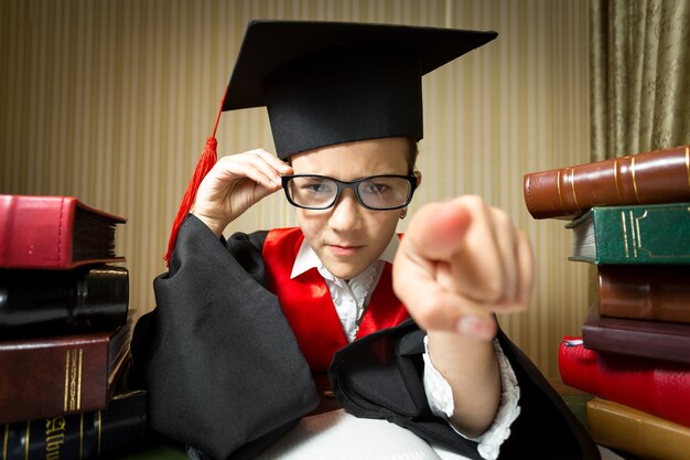 Funny portrait of girl in eyeglasses and graduation cap pointing at camera with finger