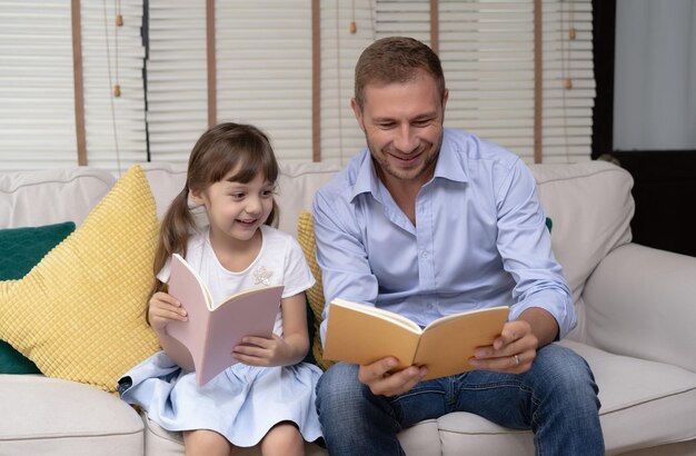 Funny portrait of smiling father and her daughter sitting sofa reading a books at homehappy father's day