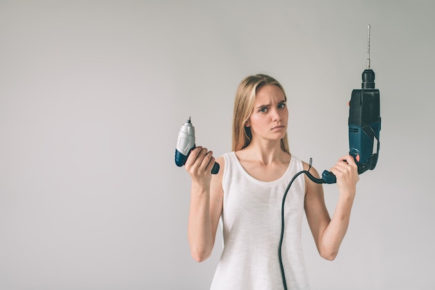 Funny portrait upset craftswoman. Blonde woman is wearing shirt isolated on white. Girl does not know how to use a drill