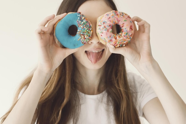 Foto ritratto divertente di una giovane modella femminile che tiene ciambelle blu e rosa vicino agli occhi e mostra una lingua