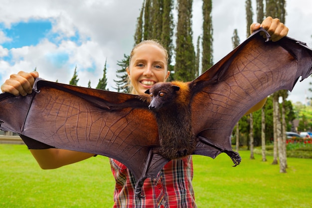 Photo funny portrait of young girl holding in hands giant flying fox ( fruit bat ) during traveling in tropical bali island.