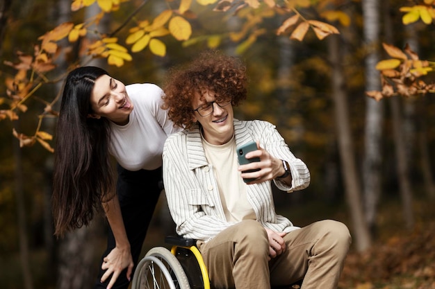 재미있는 긍정적인 장애인 남자와 그의 여자친구는 전화로 사진을 찍습니다 공원 야외에서 휠체어를 타고 웃고 있는 청년