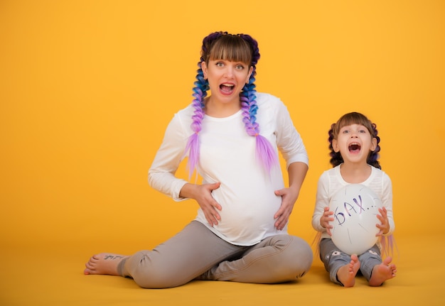 Funny positive mom and daughter show their pregnant belly posing on a yellow wall. The concept of foolishness and family play. Copyspace