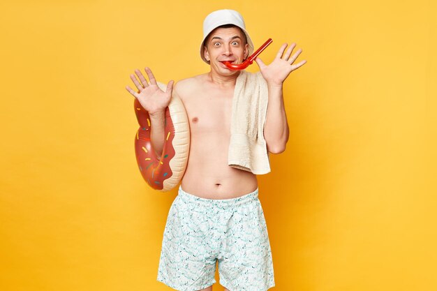 Funny positive optimistic man wearing shorts swimsuit and panama holding donut rubber ring isolated on yellow background raised arms holding snorkel in mouth