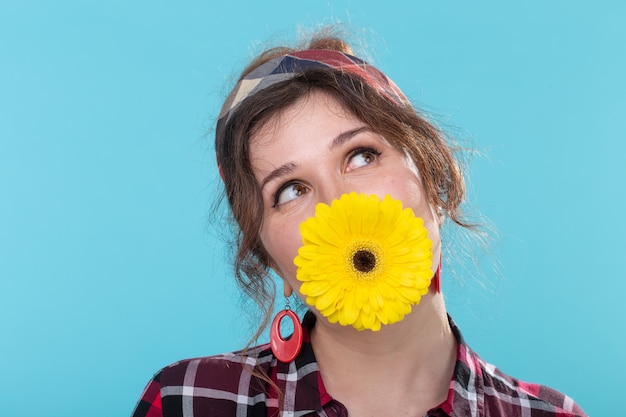 Funny positive young woman in retro image holding a bright yellow gerbera flower in her teeth posing against a blue surface