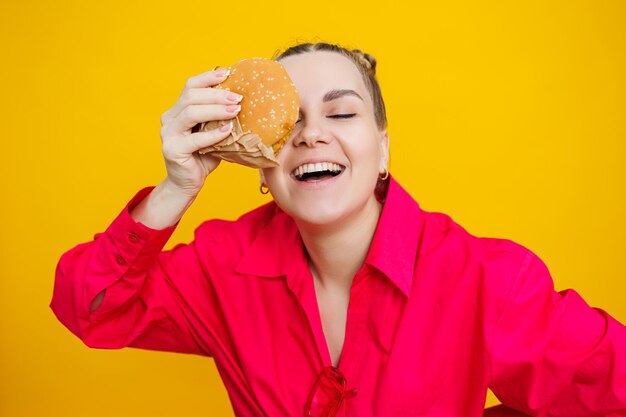 Funny pregnant young woman eating burger pregnant woman holding tasty sandwich while standing isolated on yellow studio wall junk food concept fast food during pregnancy