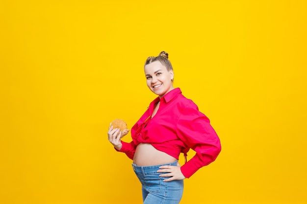 Photo funny pregnant young woman eating burger pregnant woman holding tasty sandwich while standing isolated on yellow studio wall junk food concept fast food during pregnancy