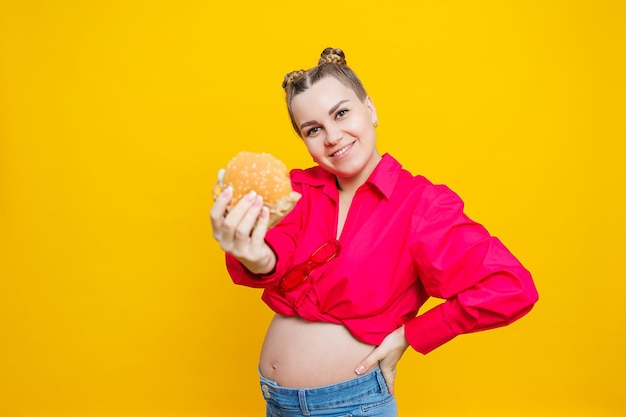 Funny pregnant young woman eating burger Pregnant woman holding tasty sandwich while standing isolated on yellow studio wall Junk food concept Fast food during pregnancy