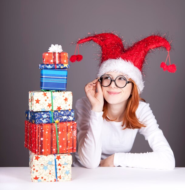 Divertente ragazza dai capelli rossi in cappello di natale e bicchieri con scatole regalo.