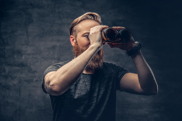 Funny redhead bearded male looking through two craft beer bottle like through binoculars.