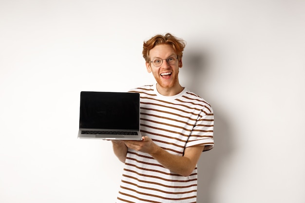 Funny redhead man in glasses showing laptop screen advertisement and smiling. Guy with red hair demonstrates promo or banner on display, white background.