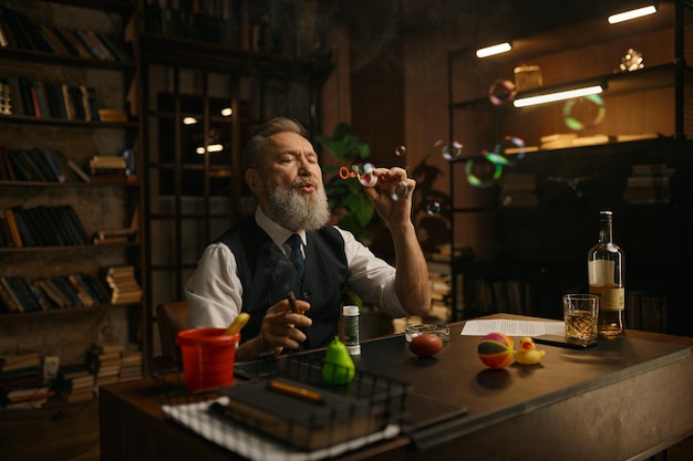 Funny relaxed senior businessman blowing up soap bubbles while sitting at work table