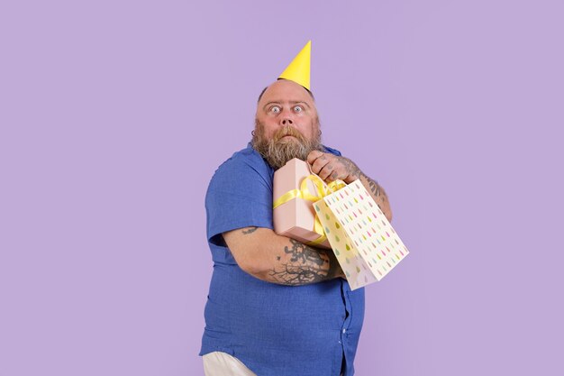 Funny scared man with overweight in party hat holds presents on purple background