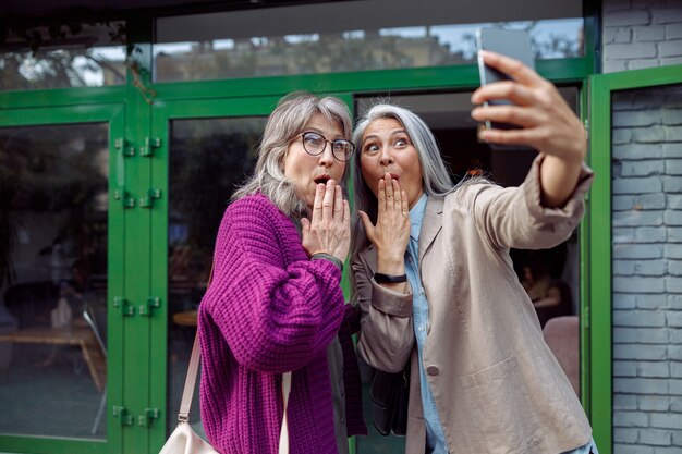 Funny senior ladies take selfie covering mouthes with palms on city street