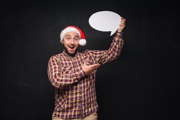 Funny serious  man in red Christmas Santa hat holds empty white cardboard as blank or mock up with copy space for text. Black background