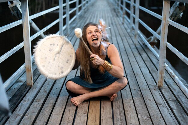 Foto femmina sciamana divertente che suona il tamburo sciamanico su un ponte di legno espressione facciale