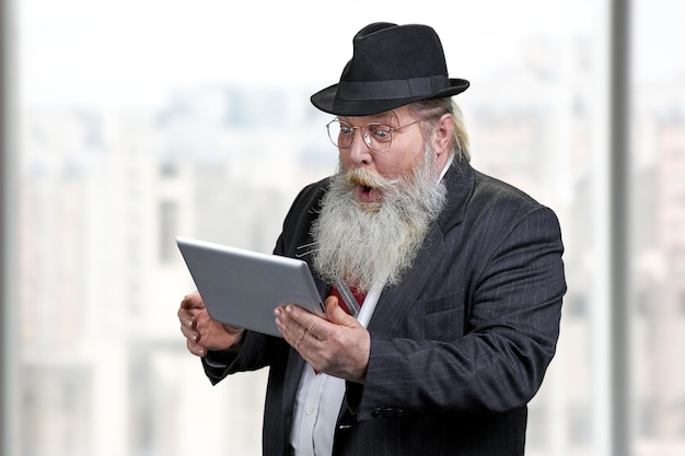 Funny shocked senior businessman using tablet pc. Blurred office window background.