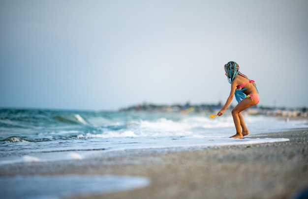 Funny slender cheerful girl playing with small waves, kicking their feet and spinning in place under the warm bright summer sun enjoying the long-awaited vacation