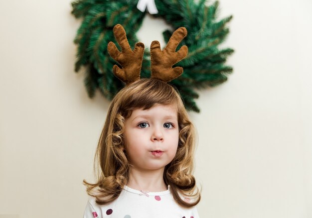 クリスマスの花輪を持つ面白い小さな女の子。ミニマリストスタイル。鹿の角を持つかわいい子供。