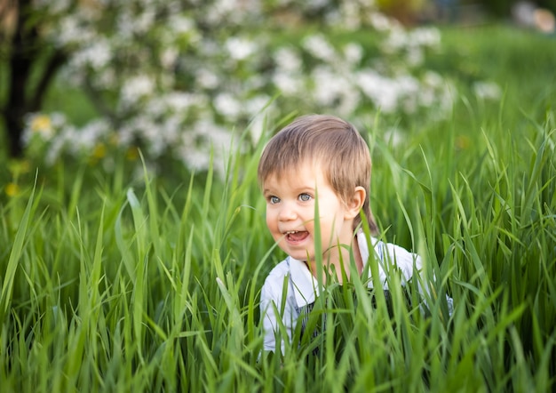 Funny smiling boy in denim blue overalls and bright blue eyes. It is funny hiding in tall green grass in a warm spring garden