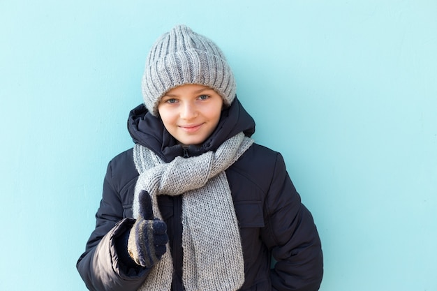 Photo funny smiling child showing thumbs up, ready for winter vacation. fashionable boy in winter gray cap and scarf standing against blue wall.