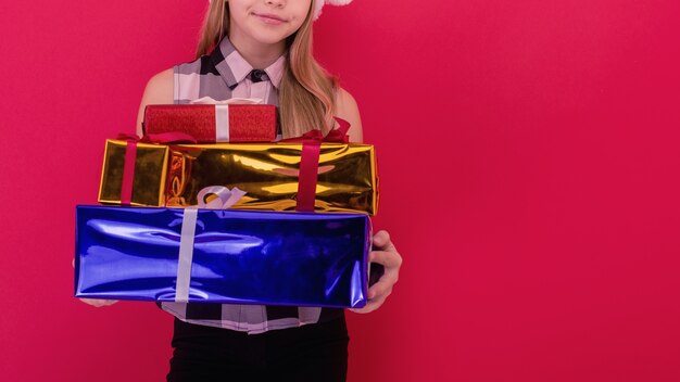 Una bambina sorridente e gioiosa con un cappello da babbo natale che tiene in mano un regalo di natale sullo sfondo rosso