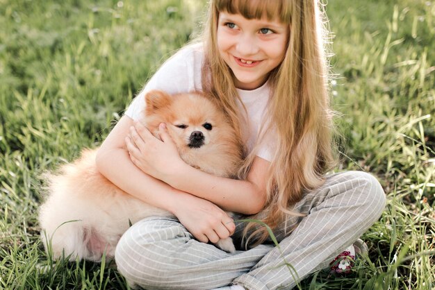 Funny smiling kid girl playing with spitz dog sitting in green grass