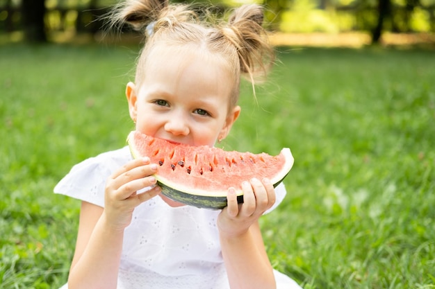 緑の芝生でスイカを食べる白いドレスの面白い笑顔の子供の女の子