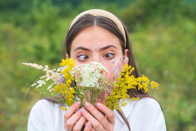 写真 面白い春の若い女性の屋外。白い花のフィールドで美しい十代の少女。楽しい顔の感情。野花の花束を持つ少女。