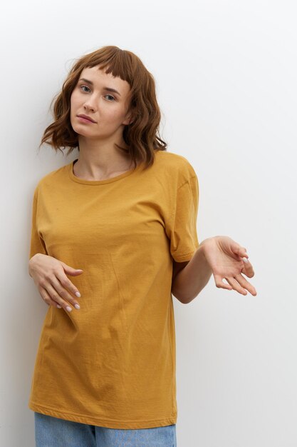 A funny surprised woman stands on a white background in a yellow cotton t-shirt and poses gesturing with her hands