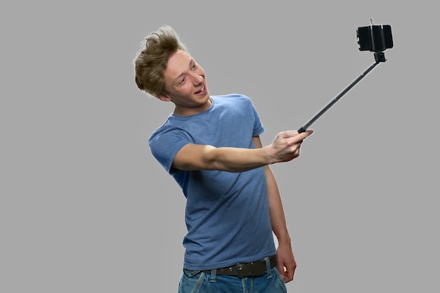 Funny teen boy using selfie stick. Stylish teen guy taking selfi with monopod against gray background. Youth and modern technology concept.