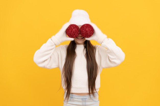 Funny teen girl ion winter hat and gloves hold decorative christmas balls on yellow background xmas