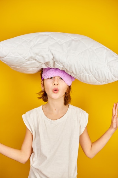 Смешная девочка-подросток в белой пижаме с фиолетовой маской для сна держит подушку