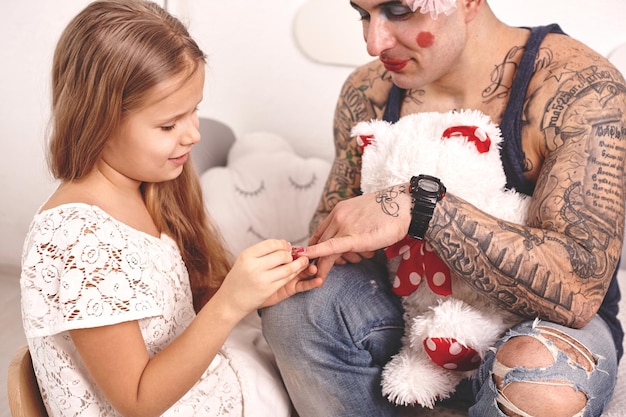 Забавное время: татуированный отец в кепке и его ребенок играют дома, милая девушка делает ему макияж ...