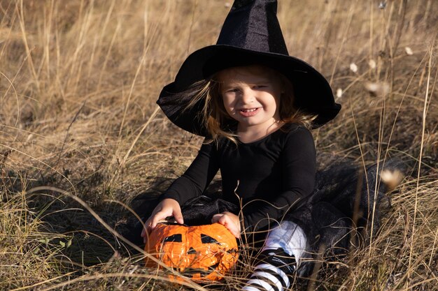 Funny toddler girl in black witch hat with orange candy bucket in nature