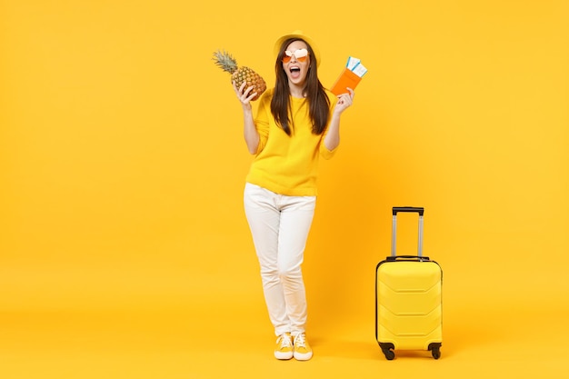 Funny traveler tourist woman in hat hold passport, tickets, fresh ripe pineapple fruit isolated on yellow orange background. Passenger traveling abroad on weekends getaway. Air flight journey concept.