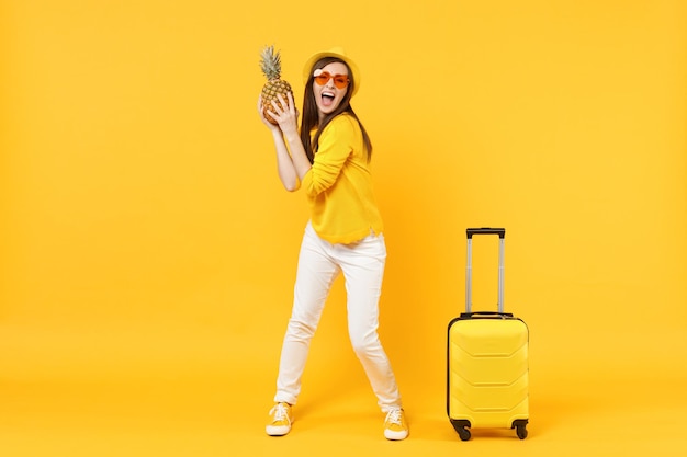 Funny traveler tourist woman in summer clothes, hat holding fresh ripe pineapple fruit isolated on yellow orange background. Passenger traveling abroad on weekends getaway. Air flight journey concept.