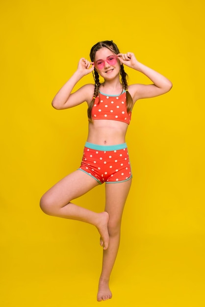Забавная девочка в подростковом возрасте смотрит в камеру, позируя на одной ноге на ярко-желтом фоне