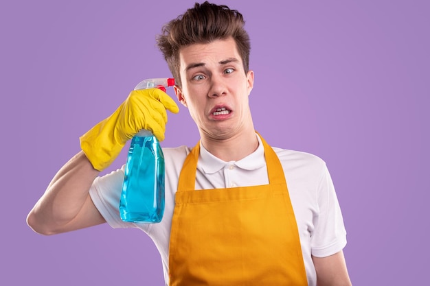 Funny unhappy young man in yellow apron and rubber gloves holding plastic bottle, with disinfectant spray near temple like gun while being tired from cleaning work during coronavirus pandemic