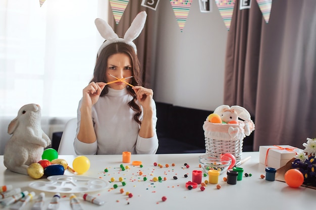 Funny upset young woman prepare for Easter alone. She sit at table in room and hold brushes crossed. Model look on camera. Decoration with paint and sweets on table.