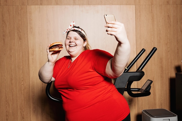 funny woman dressed in sportswear and with a bandage on her head is making selfie standing with burger in her hand next to the treadmill