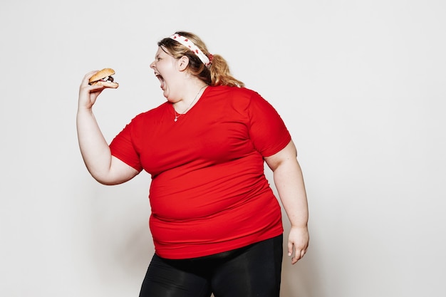 funny woman dressed in sportswear and with a bandage on her head is standing with a burger in her hand against a white wall