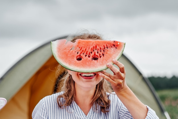 Funny woman eating watermelon on a picnic. girl closed her eyes with a watermelon, looking into the holes