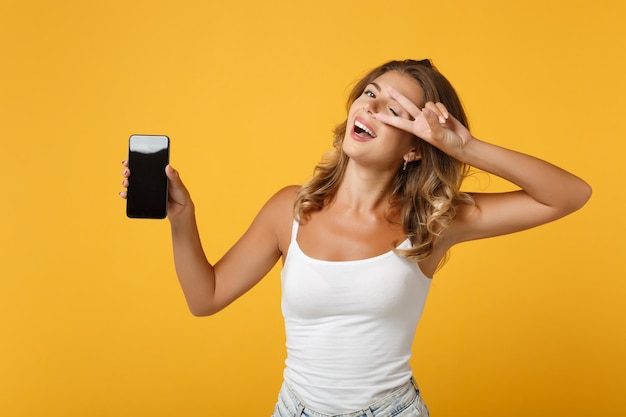 Funny woman girl in light casual clothes posing isolated on yellow orange background. people lifestyle concept. mock up copy space. holding mobile phone with blank empty screen, showing victory sign