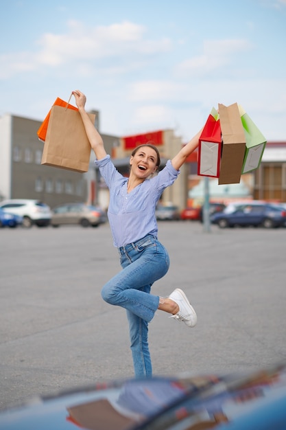 面白い女性はスーパーマーケットの駐車場で段ボールの袋を持ってジャンプします。ショッピングセンターからの購入、バックグラウンドでの車両を運ぶ幸せな顧客