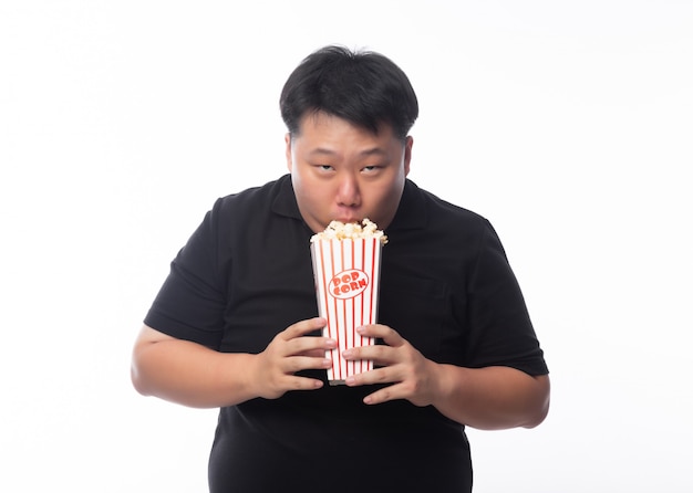 Смешной молодой азиатский толстый человек в черной рубашке поло ест попкорн на белом фоне
