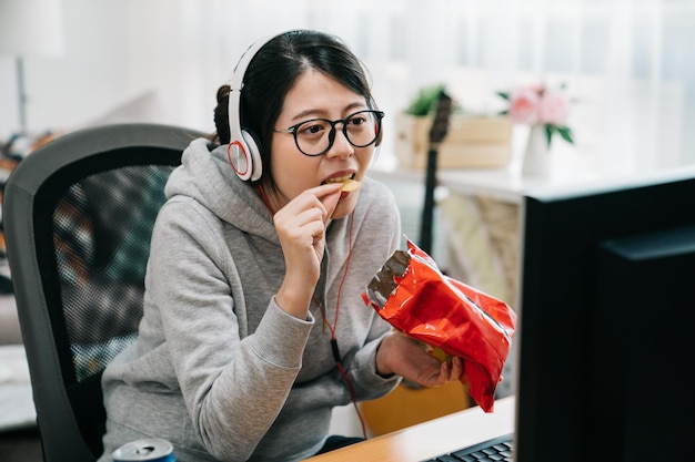 재미있는 젊은 아시아 여성 오타쿠는 칩을 먹고 컴퓨터 화면에서 비디오를 보는 손에 간식 팩을 들고 집에 앉아 있습니다. 여름 방학에 여학생이 정크 푸드로 인터넷을 봅니다.