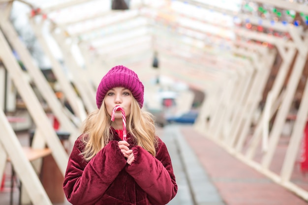 Смешная молодая блондинка в красной вязаной шапке позирует с леденцом на улице