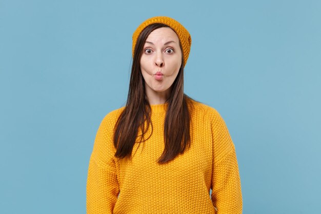 Funny young brunette woman girl in yellow sweater and hat posing isolated on blue wall background studio portrait. People sincere emotions lifestyle concept. Mock up copy space. Blowing cheeks lips.